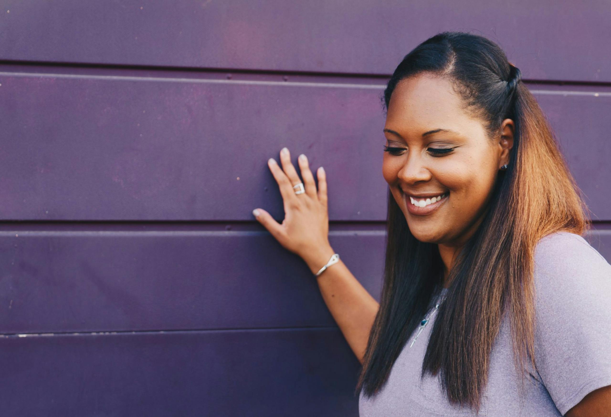 A young woman smiles against a purple wall, exuding happiness and style.