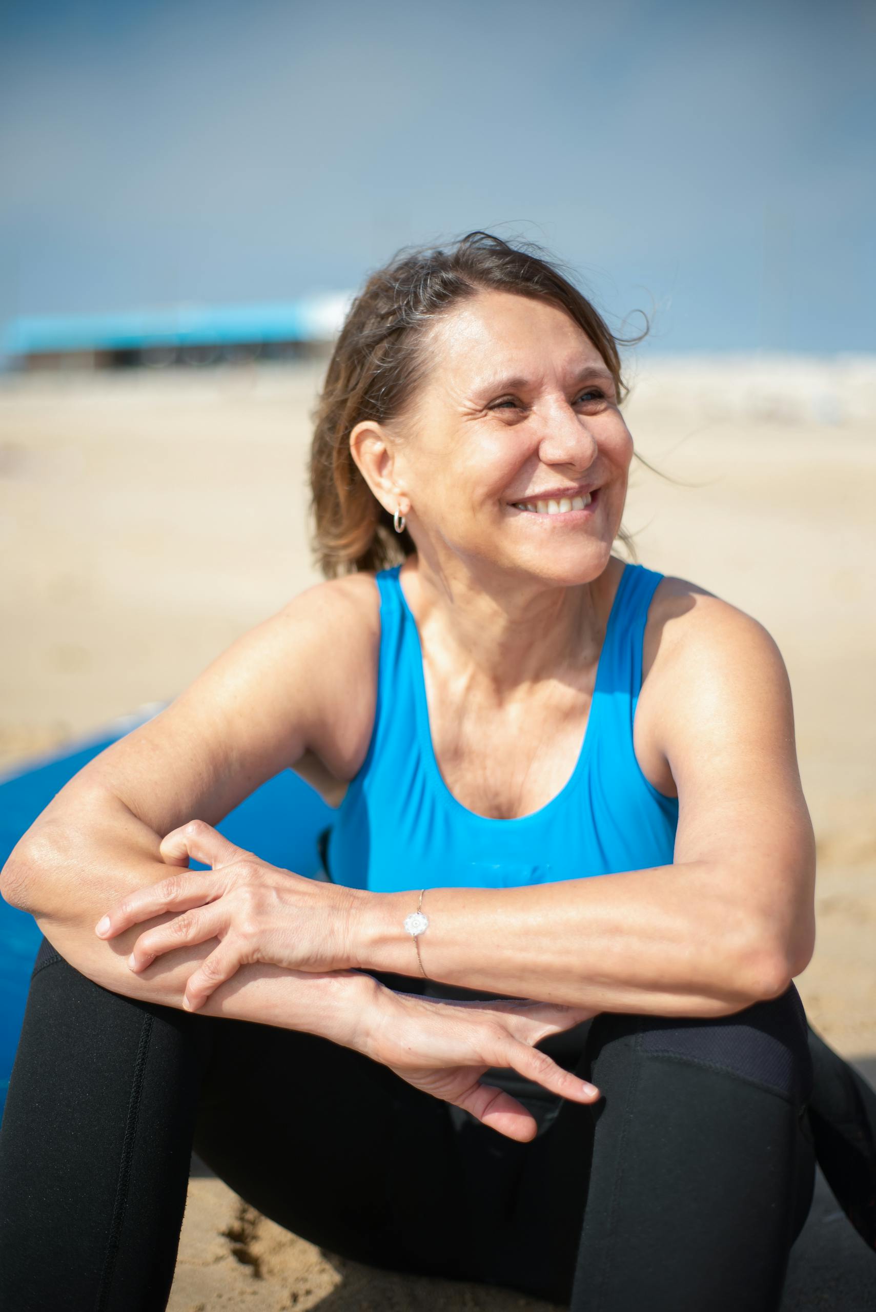 Senior woman wearing blue tank top, smiling and sitting on the beach, enjoying a sunny day outdoors in Portugal.